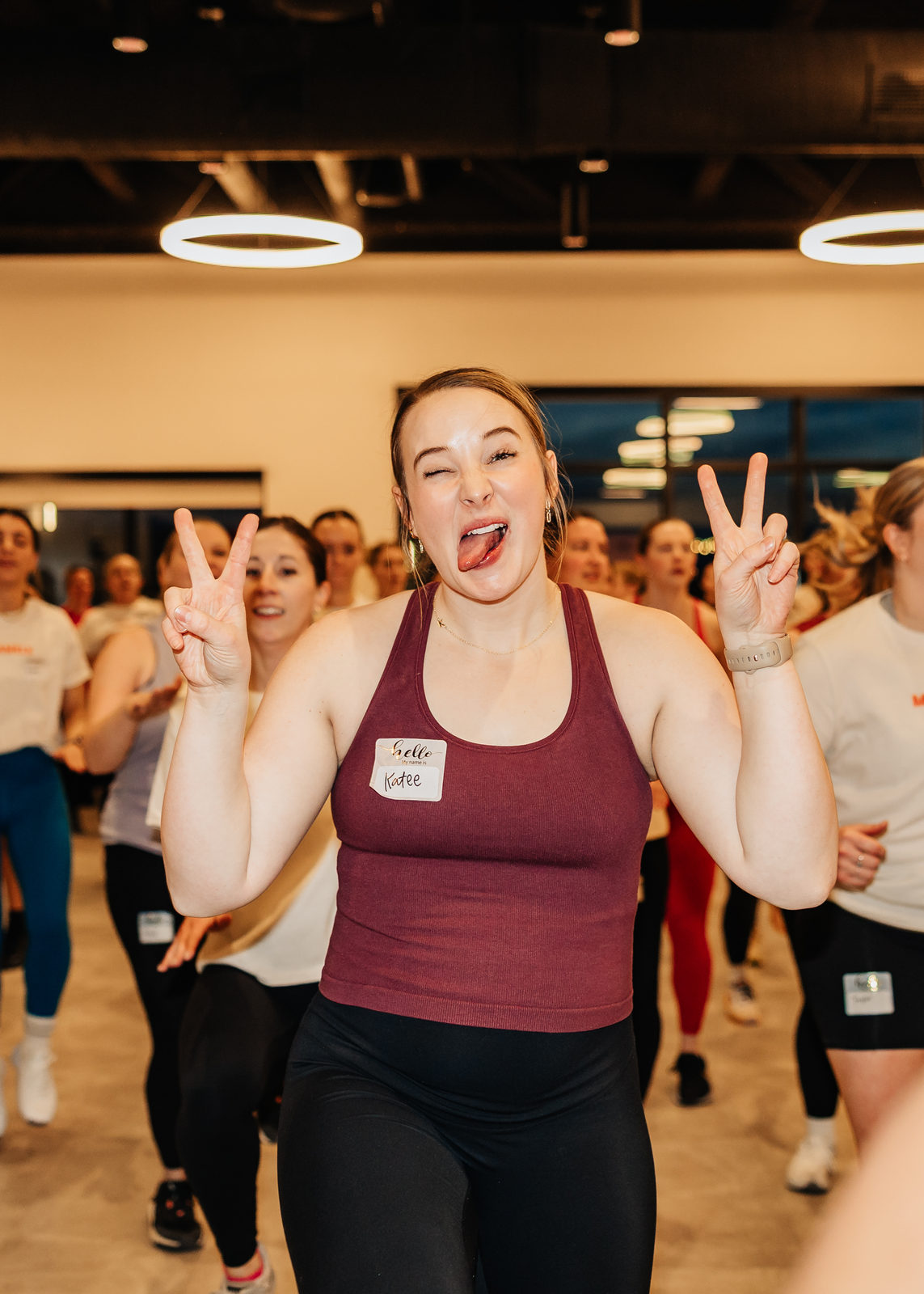 A first-time member with her name tag on, grinning and throwing peace signs mid-workout in a packed studio.