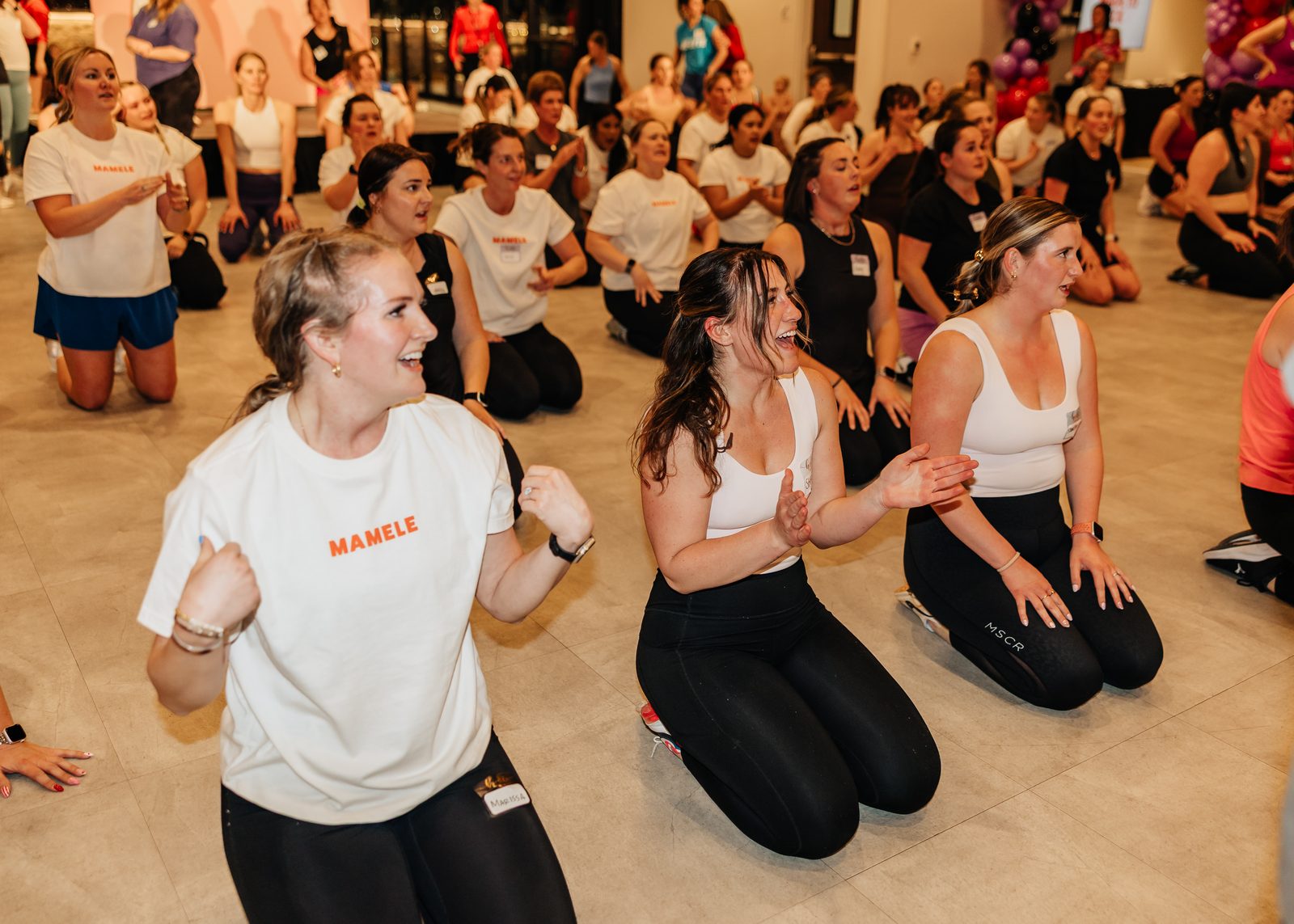 Two members kneeling and laughing together partway through a group workout, the rest of the room behind them.