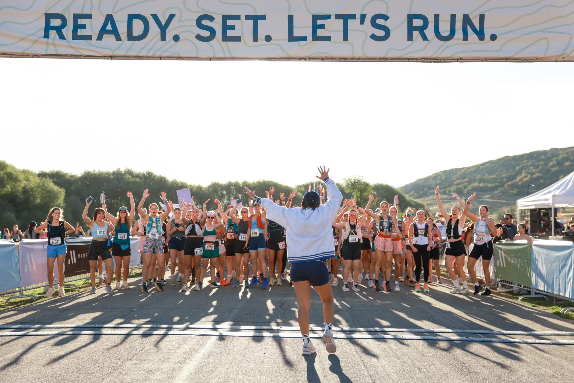 The 2026 Mamele Mountains start line: dozens of women with bibs cheering with arms up under the Ready Set Let's Run banner.