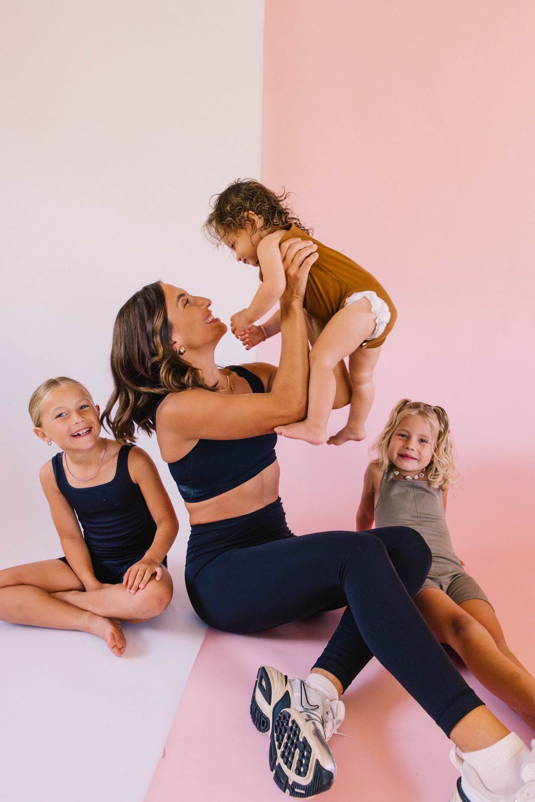 A mom in workout clothes lifting her baby on the floor, her older daughters in frame.