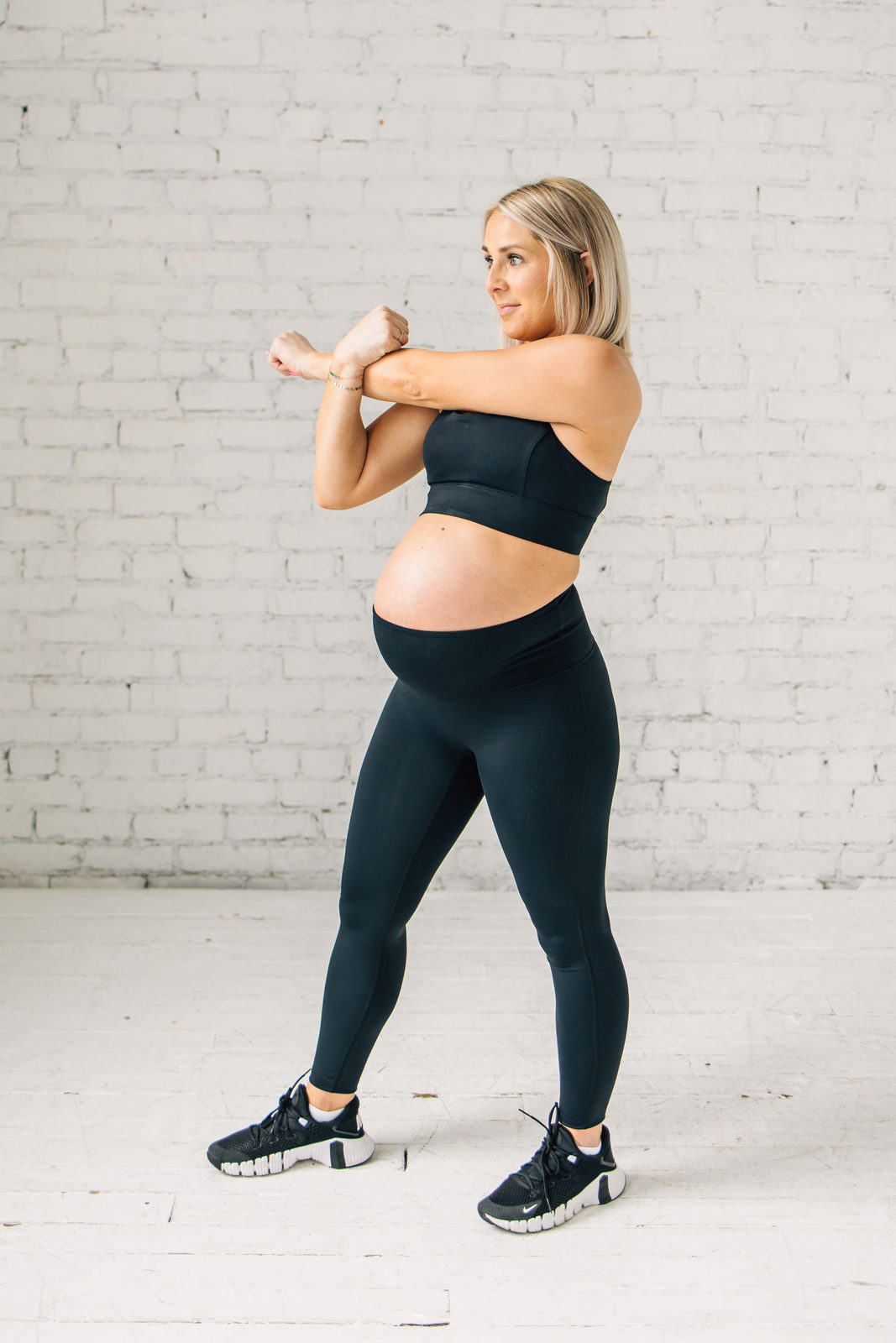 A visibly pregnant woman mid arm-swing in Mamele athletic wear against a white brick wall.